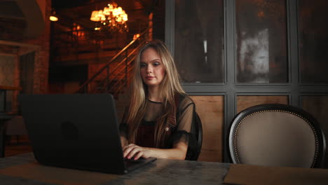 Young-woman-sitting-in-coffee-shop-at-wooden-table,-drinking-coffee-and-using-smartphone.On-table-is-laptop.-Girl-browsing-internet,-chatting,-blogging.-Female-holding-phone-and-looking-on-his-screen.